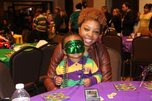 The photo shows a woman with a child in a Mardi Gras mask sitting at the St. Ignatius Loyola Catholic Church event.