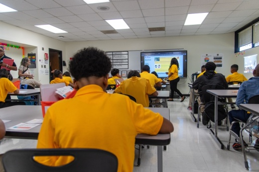 students sit in desks facing toward the front of a classroom where a teacher walks in front of a screen
