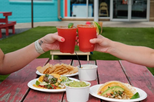 Two arms reach across a picnic table holding tacos and cheers each other with red-colored margaritas.