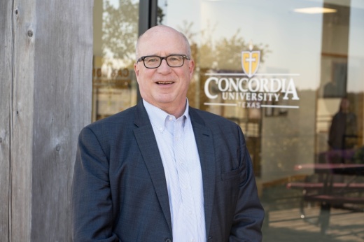 A man with glasses in a suit smiles in front of a building that reads Concordia Univeristy