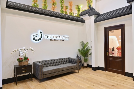 a room with a grey couch, side table with flowers on top and a sign above it that says "The Total You Medical Spa"