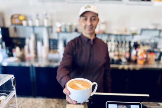 The owner holds a white cup with coffee inside while standing behind the counter.