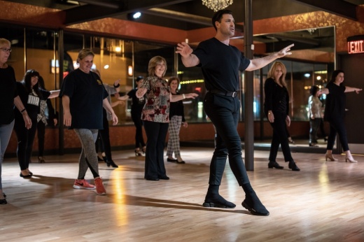 A male teacher in all black shows a group of adults behind him how to dance