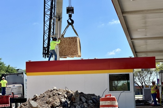 A construction worker standing on top of the kiosk helping a crane guide a wooden box onto the roof with his hands.