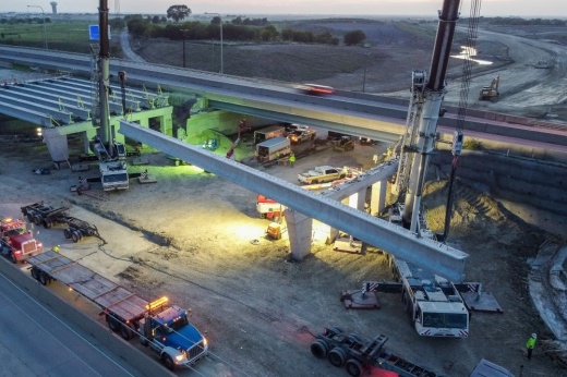 Construction crews lift a beam up onto a bridge deck that crosses Fields Parkway.
