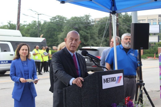 a man stands a a podium with a microphone with a street and traffic in the background
