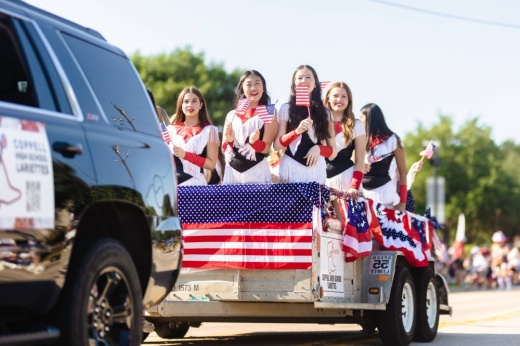 Females wearing red, white and blue holding American flags riding on a trailer in a parade.