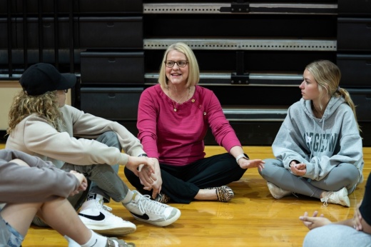 woman sits on ground with teenagers in a gymnasium