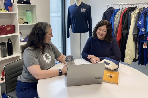 two women sit at a table talking and looking at a laptop
