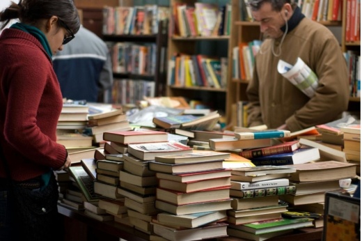 two people look over piles of books on a table in a store