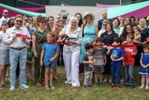 A group of children and adults stand behind the ribbon as director Cheryl Kruckenberg cuts the ribbon.
