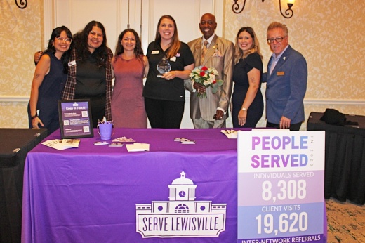 Group of males and females standing behind a table with Serve Lewisville table cover.