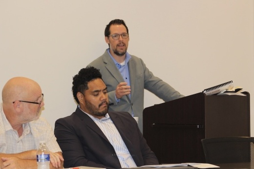Picture of a man talking at a podium while two seated men listen.