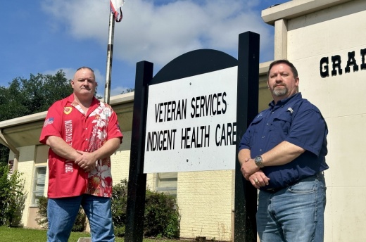 The two men stand side by side and pose for a photo in front of the officer's sign and flagpole.