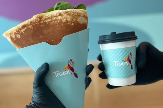 Two hands hold a crepe and a coffee in Tecuen-branded packaging.
