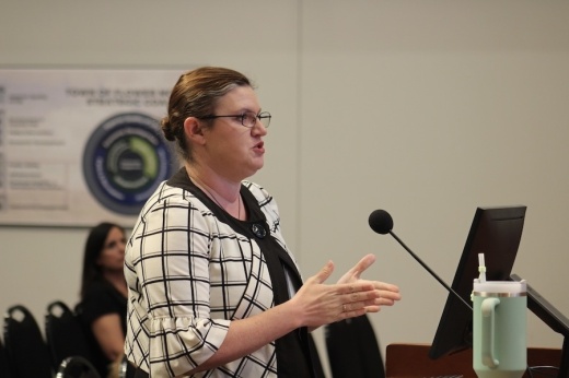 Picture of a woman standing at a lectern and talking with council members.