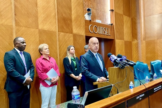 a man in a suit stands at a dais with several microphones and four people behind him