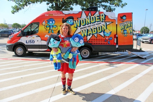 Female wearing super hero costume holding super hero stuffed objects in front of a van with Wonder Wagon signage.