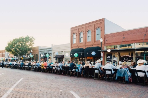 Group of males and females sitting along a long table in the middle of street lined with old buildings.