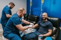 One man sitting in a chair while another man prepares his arm for an IV injection.
