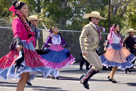 The dancers are shown with the women dressed in colorful clothing with flowing skirts and men dressed in beige suits with matching hats.