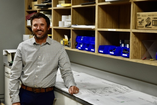 Posed photo shows Randall Malik leaning on a desk with papers on it next to a large array of shelves on the wall.