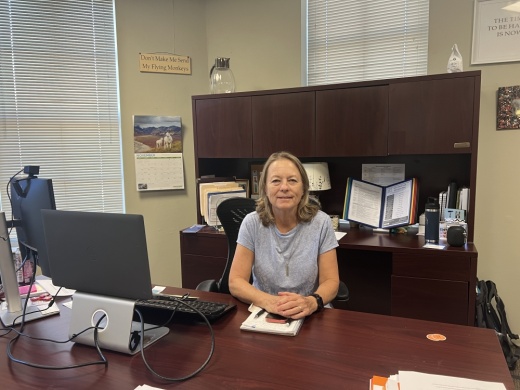 A white woman with collar-bone length blonde hair sits at a brown desk with her hands crossed in front of her.