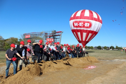 Males and females using ceremonial shovels to turn dirt wearing red hard hats. An oversized H-E-B grocery cart and hot air balloon are also pictured.