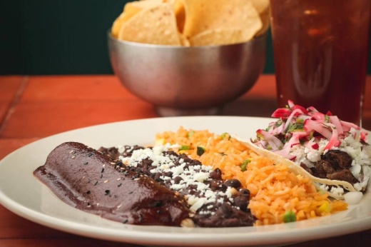Plate with two enchiladas and side of rice and beans with bowl of chips in background.
