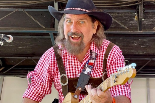 The musician smiles while playing guitar and wearing a black cowboy hat and red and white checkered shirt during a previous show.