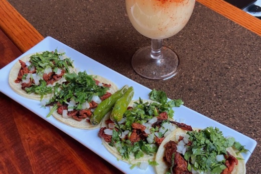 A street taco plate is shown filled with toppings like cilantro, onions and roasted whole peppers. Part of a margarita is also shown.
