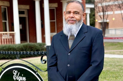 The interim city manager stands outside of city hall and smiles for a photo wearing a suit and tie.