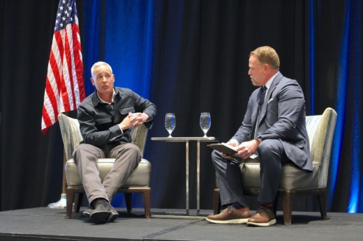 Two men sit in chairs on a stage.