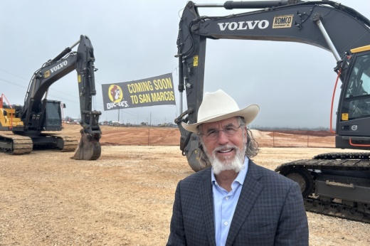 A man with a cowboy hat stands in front of two construction machine that hold a sign that says "Coming Home to San Marcos"