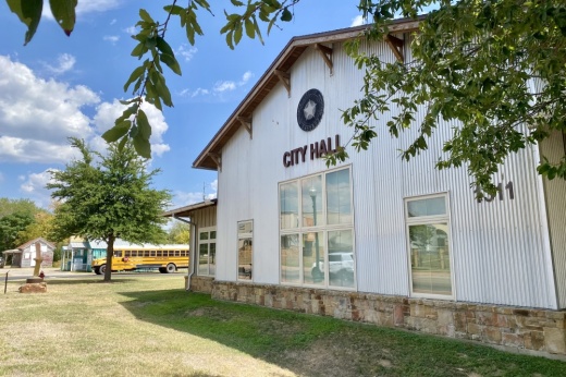 The white exterior of city hall is shown with a blue sky with some white clouds and green grass and leaves.