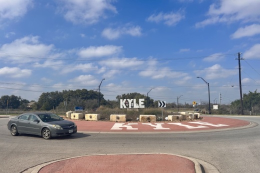 A sign that says Kyle in a roundabout with a car in front of it.