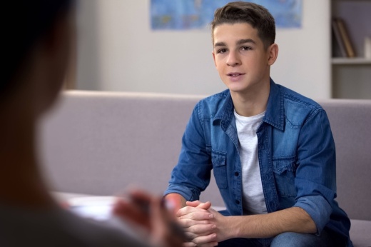 Male teen sitting on a couch facing another seated person with back to the camera.