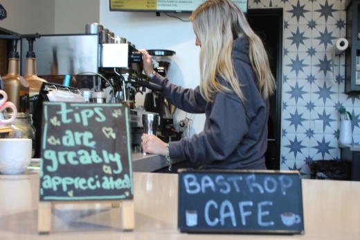 A barista makes coffee in the background in the camera's focus, while two tiny chalkboard signs, reading Bastrop Cafe and Tips are greatly appreciated, are blurred in the foreground.