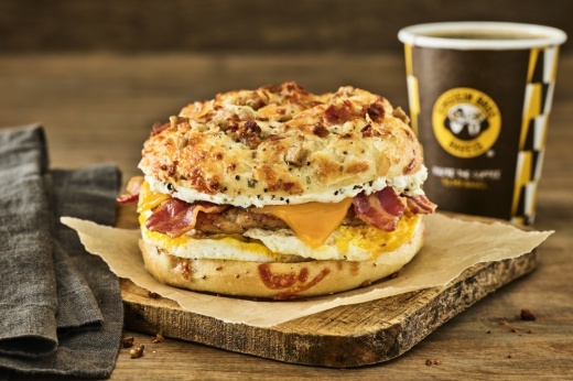 A wooden slab holds an everything bagel with eggs, cheese and bacon, with a coffee in the background.