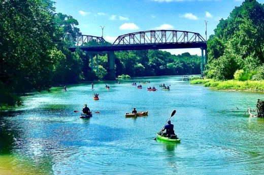 People paddle down the blue river with green trees on the bank and the Old Iron Bridge in the background.