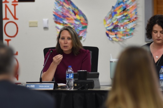 Trustee Trish Bode is framed by out of focus heads as she gestures while talking from the dais.