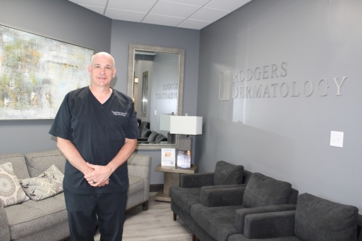 Male wearing dark medical scrubs standing in an office lobby with Rodgers Dermatology signage.