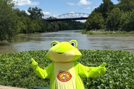 Jack Plimpton wears a frog costume—the company's mascot—and gives two thumbs-up in front of Bastrop's Old Iron Bridge and the Colorado River.