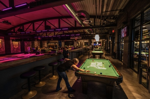 A woman in a black t-shirt and pants lined up to take a shot in a game of pool, in the back is a large bar with wood floors and pink neon lighting.