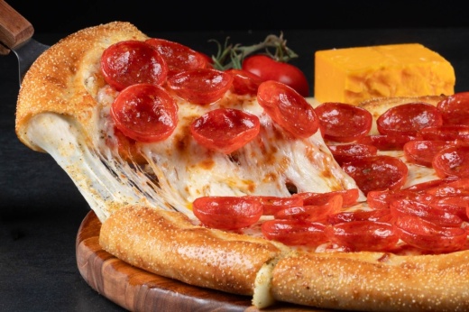 Slice of pepperoni pizza being pulled away from the pizza pie while cheese stretches between the slice and the pie.
