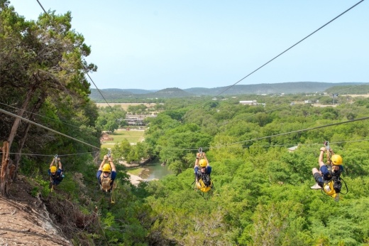 4 people ride on a zip line high above the tree line on a sunny day in the Hill Country.