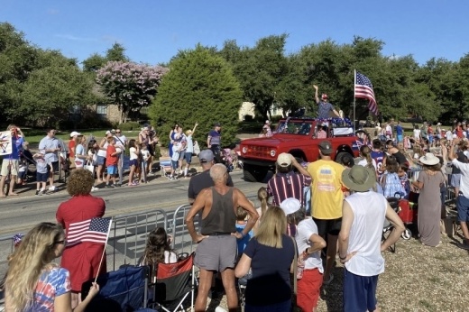 Image of a Fourth of July parade where a red truck is driving by as a float with people on it and flying an American flag. Onlookers watch with flags and festive attire.