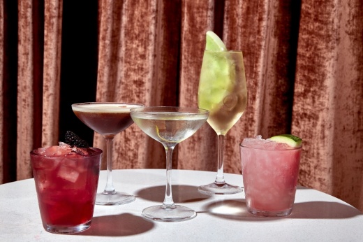 Images shows five cocktails on a table with a white tablecloth and an orange velvet backdrop. From left to right to right is a short red cocktail on the rocks either a blackberry garnish, a brown martini, a clear martini, a full champagne flute with a green melon garnish and a short pink cocktail with a watermelon garnish.