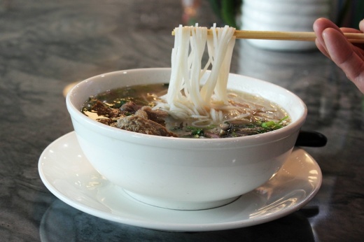 Bowl of pho soup with a person's hand holding chopsticks while pulling noodles from the bowl.