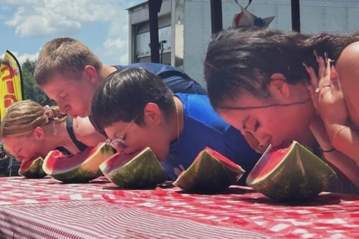 People lined up for a watermelon competition eating a slice of watermelon without using their hands.